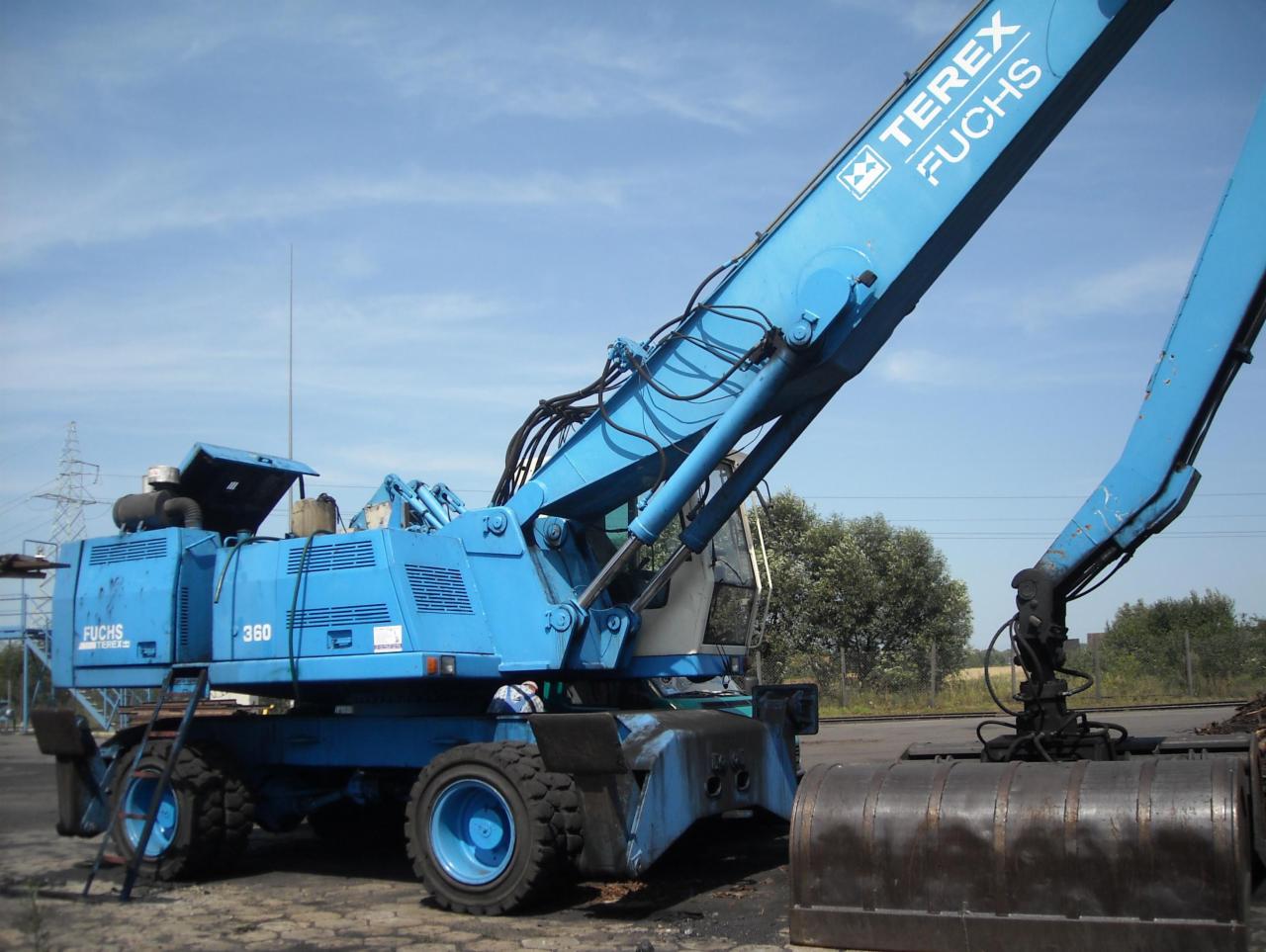 Niebieska koparko-ładowarka przemysłowa Terex Fuchs MHL 360 na placu budowy, widok z boku, z łyżką skierowaną w dół, na tle błękitnego nieba.