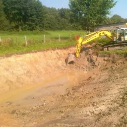 Żółta koparka Komatsu przy wykopanym stawie, w tle budynek gospodarczy i drzewa.