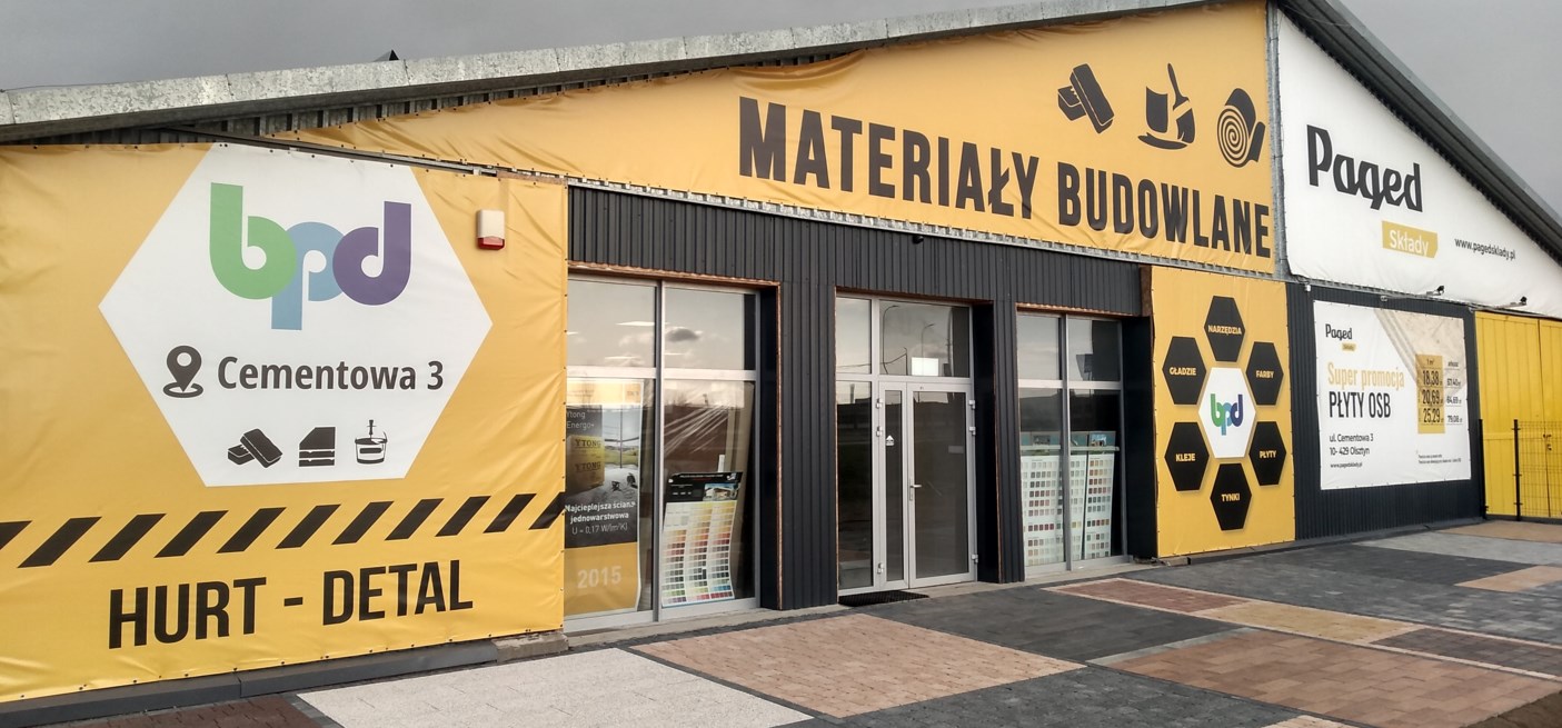 Fasada budynku składu budowlanego z dużymi banerami reklamowymi, prezentującymi logo firmy, materiały budowlane i promocje na płyty OSB.
