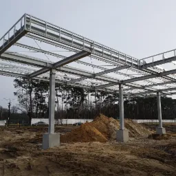 Szkielet stalowej konstrukcji z betonowymi podstawami w trakcie budowy na placu budowy z hałdami piasku i lasem w tle, widoczne elementy zespawane.