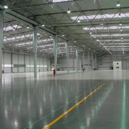 Wnętrze nowej, przestronnej hali z szarą, połyskującą posadzką, widocznymi żółtymi liniami, stalową konstrukcją dachu i licznymi lampami.