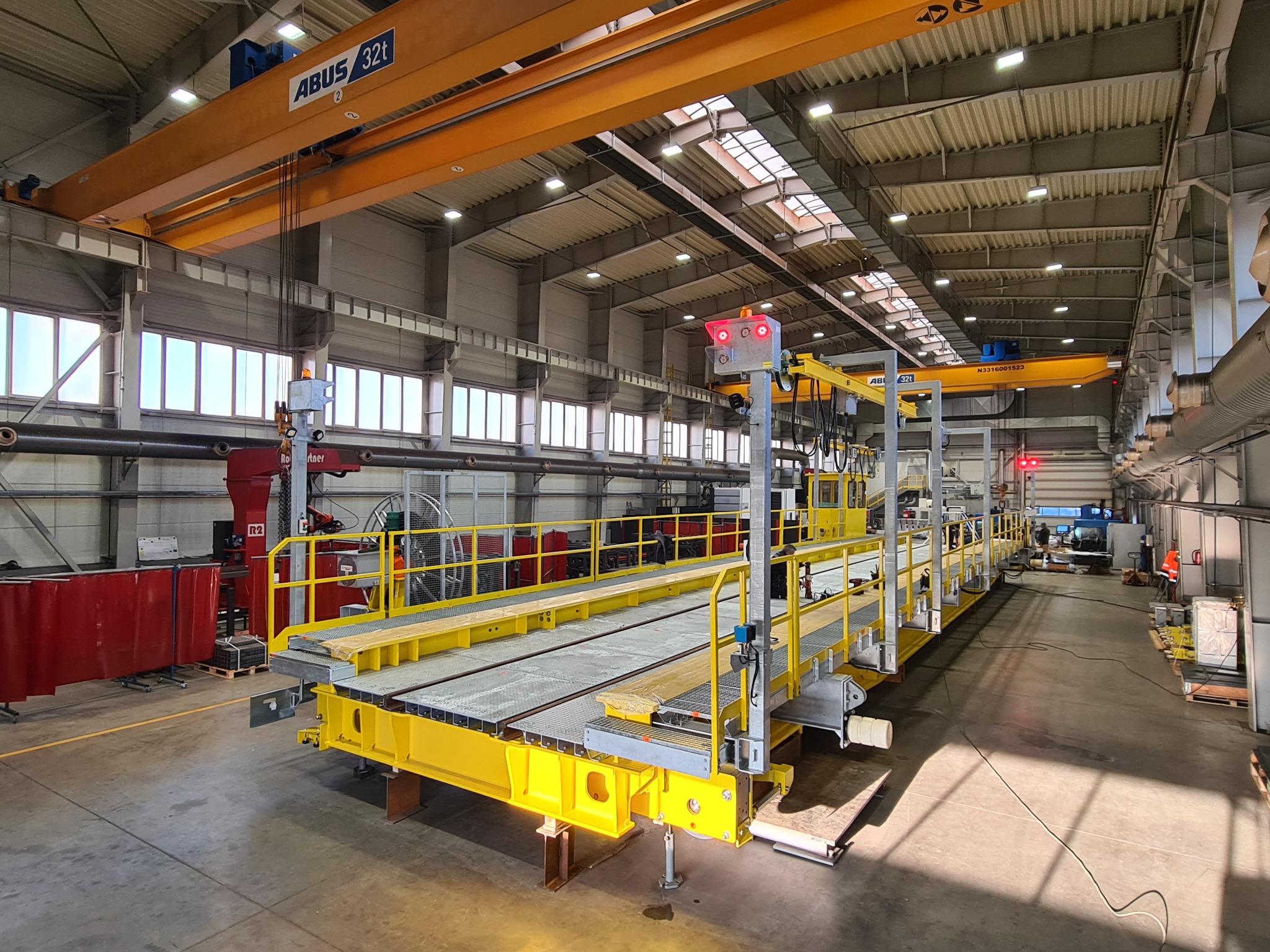 Wnętrze hali z suwnicą ABUS 32t i żółtą platformą roboczą na szynach. Widoczne okna, oświetlenie i elementy konstrukcji stalowej. Perspektywa z poziomu podłogi.
