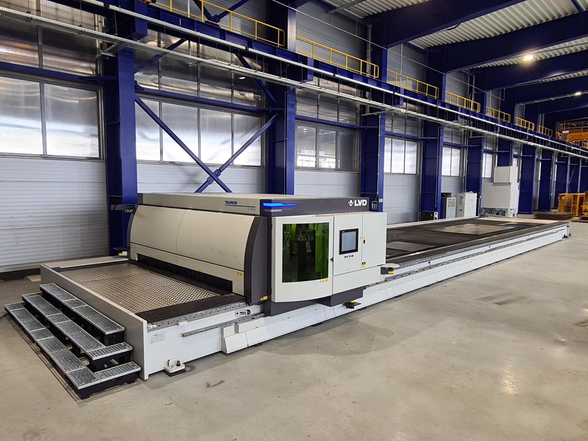 Wnętrze hali produkcyjnej z nowoczesną, biało-szarą maszyną do cięcia laserowego LVD Taurus z zielonym oknem, na tle niebieskiej konstrukcji stalowej hali.