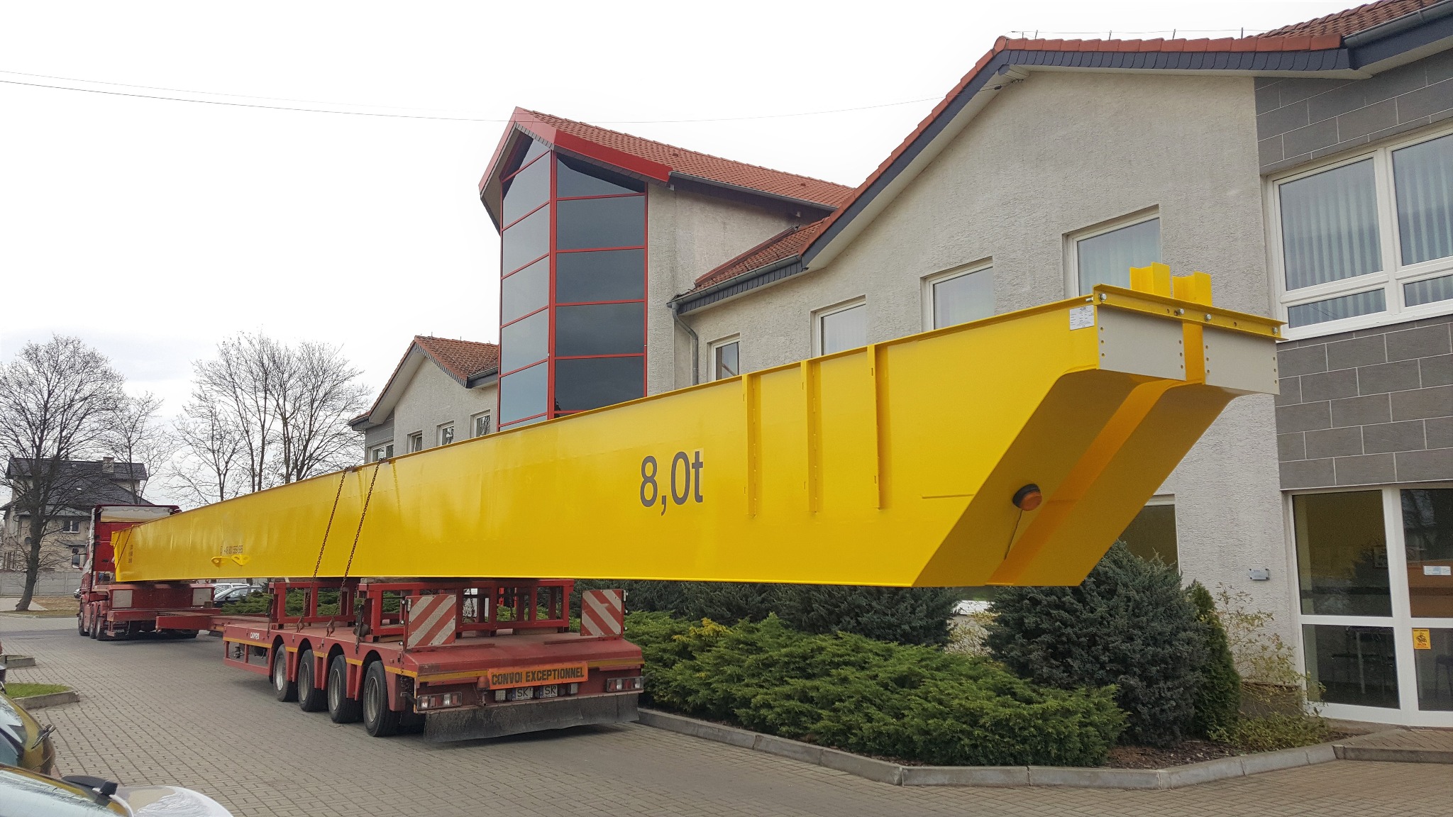 Żółty dźwig o udźwigu 8 ton transportowany na czerwonej platformie niskopodwoziowej na tle budynku. Widoczne detale konstrukcji i tablica informacyjna.