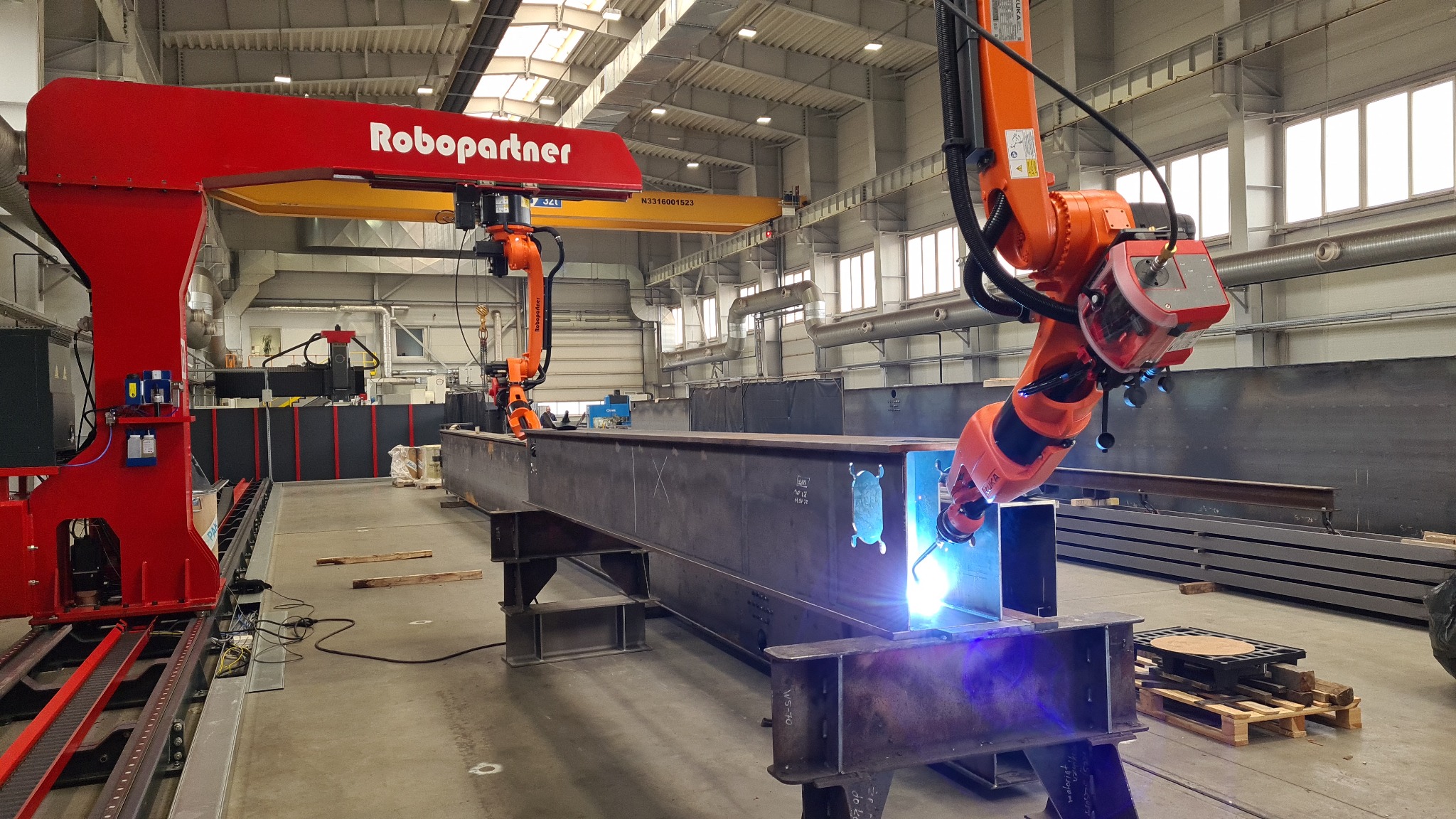 Zautomatyzowane spawanie konstrukcji stalowej przez robota Robopartner w hali produkcyjnej. Widoczny proces spawania i elementy konstrukcji.