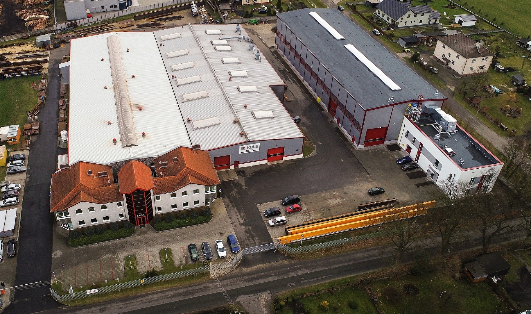 Widok z lotu ptaka na kompleks budynków przemysłowych z biurem, parkingiem i elementami konstrukcji stalowych na zewnątrz. Budynki o różnej architekturze i kolorystyce.