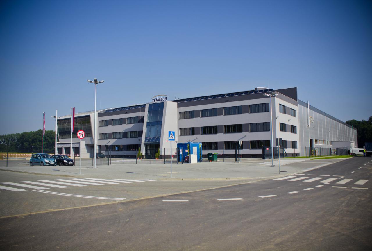 Nowoczesny budynek przemysłowy z jasnoszarą elewacją, dużymi oknami i przeszklonym wejściem, z widocznym logo firmy Tenneco na fasadzie, na tle błękitnego nieba i z elementami infrastruktury...