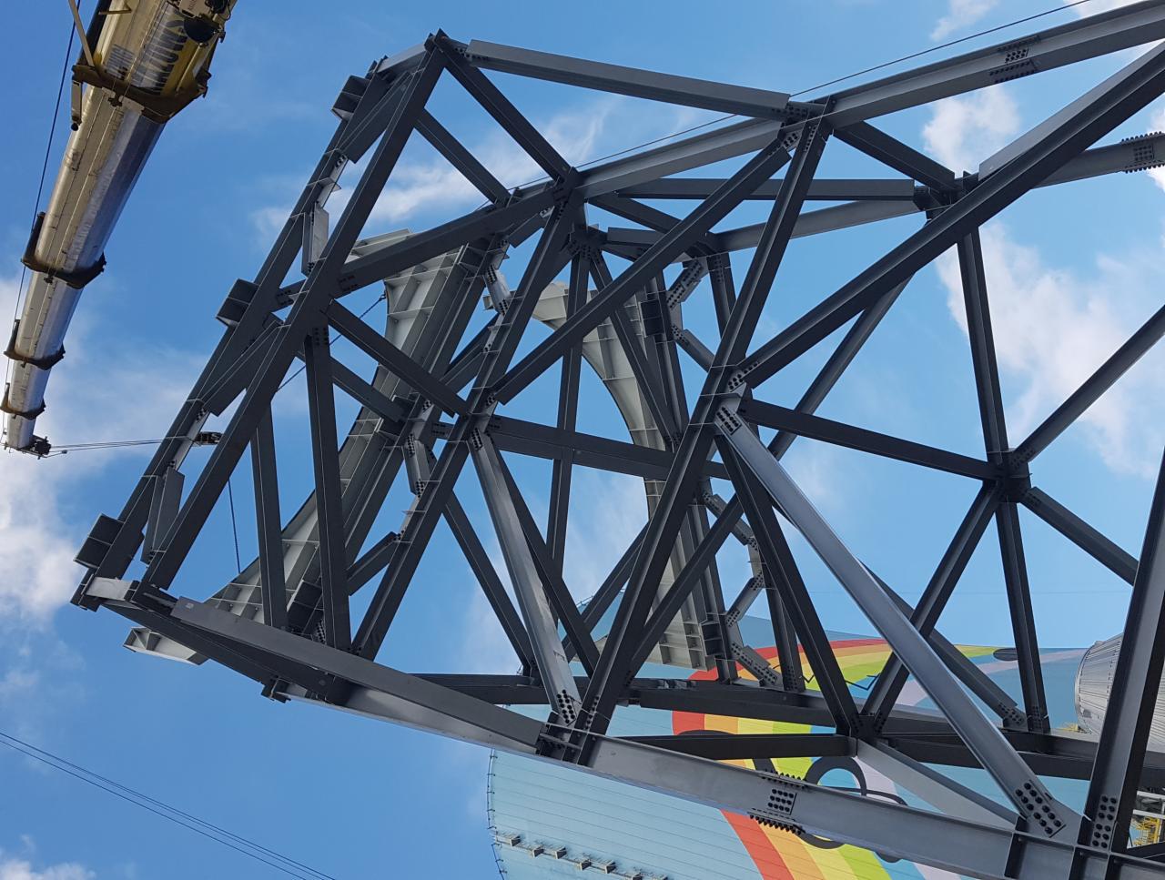 Wznoszona konstrukcja stalowa z widocznym fragmentem dźwigu, na tle błękitnego nieba i kolorowej ściany budynku.