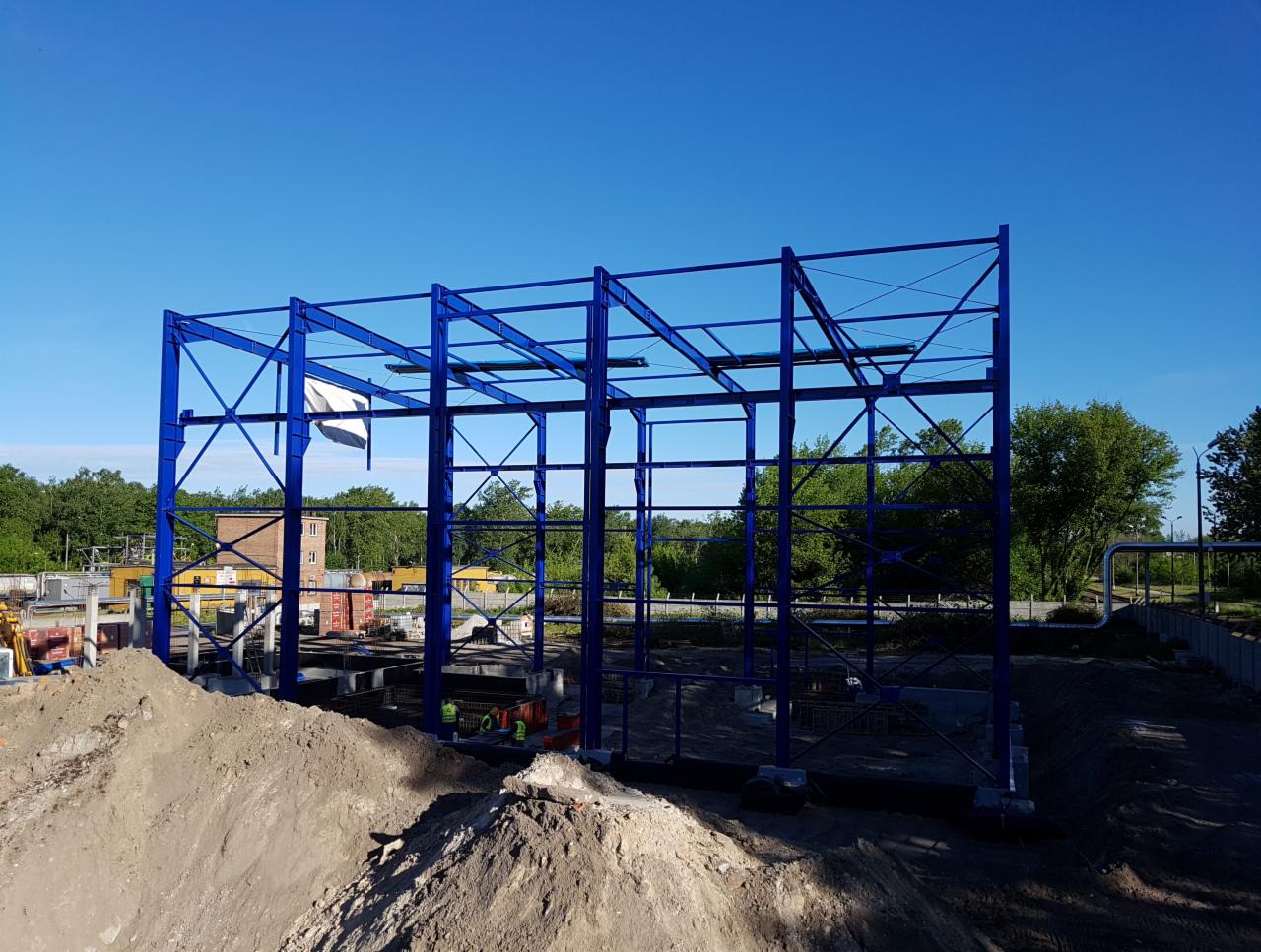 Niebieska, stalowa konstrukcja szkieletowa hali na fundamencie w trakcie budowy, widoczna sterta ziemi i elementy budowlane w tle, błękitne niebo.