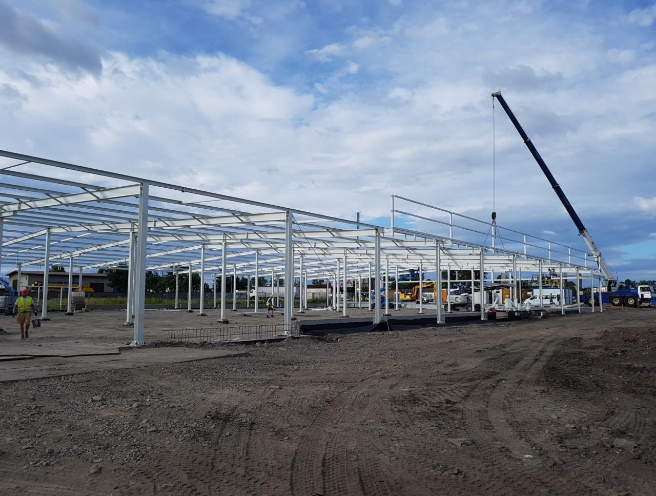 Montaż szkieletu hali stalowej w Kolonowskim. Widoczny dźwig podnoszący element konstrukcyjny. W tle błękitne niebo z chmurami.