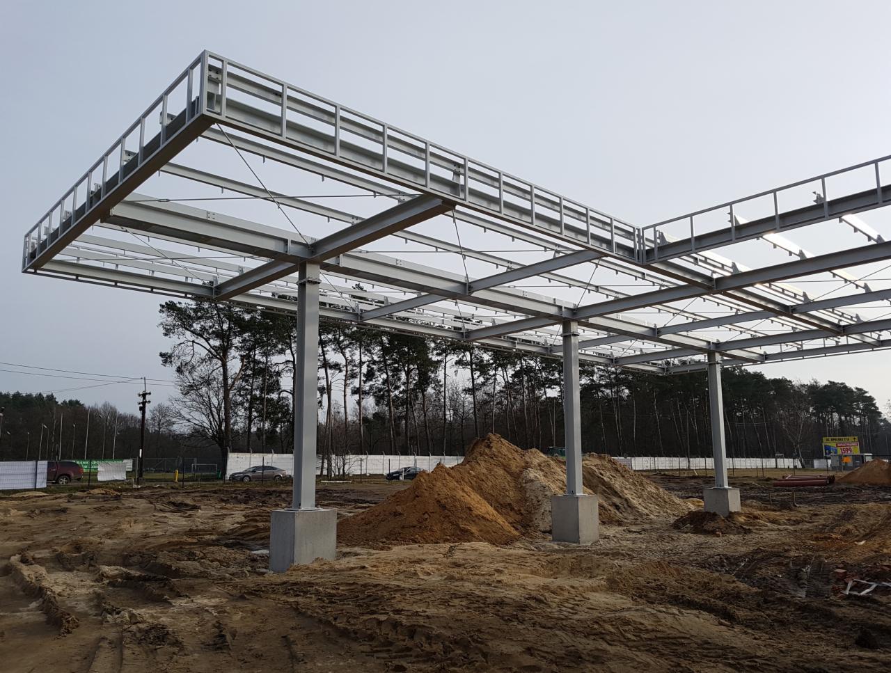 Szkielet stalowej konstrukcji z betonowymi podstawami w trakcie budowy na placu budowy z hałdami piasku i lasem w tle, widoczne elementy zespawane.