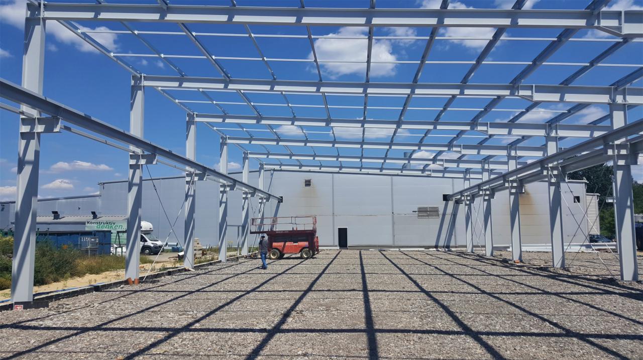 Wznoszenie szkieletu stalowego hali produkcyjnej, widoczny pracownik obok podnośnika koszowego, jasne niebo w tle.