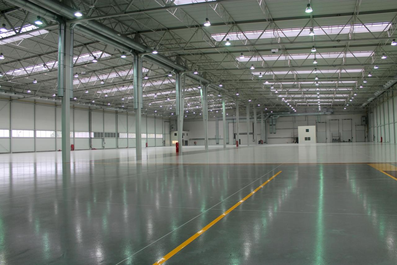Wnętrze nowej, przestronnej hali z szarą, połyskującą posadzką, widocznymi żółtymi liniami, stalową konstrukcją dachu i licznymi lampami.