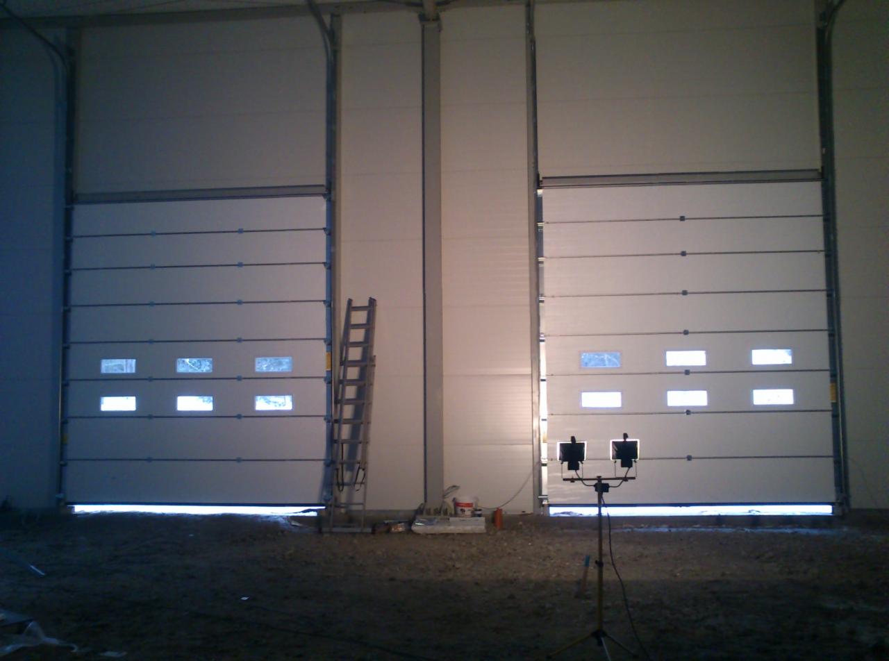 Dwie białe bramy przemysłowe segmentowe, częściowo otwarte, z oknami, oświetlone od wewnątrz. Obok drabina i lampa na statywie. Podłoże wewnątrz budynku.