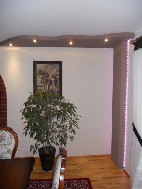 Salon po remoncie z wykończonymi ścianami, dekoracyjnym oświetleniem sufitowym, obrazem nad rośliną doniczkową i podświetlaną listwą LED przy zasłonie.