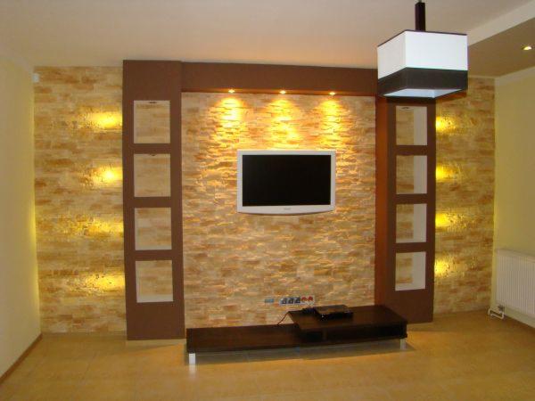 Ściana akcentująca w salonie wykończona kamieniem dekoracyjnym w odcieniach beżu i brązu, podświetlona punktowymi lampami, z wbudowanym telewizorem i brązowymi półkami po bokach, oraz wiszącą lampą...