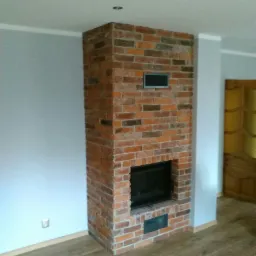 Murowany kominek z cegły klinkierowej w surowym wnętrzu, widoczna kratka wentylacyjna i wkład kominkowy, na podłodze panele imitujące drewno.