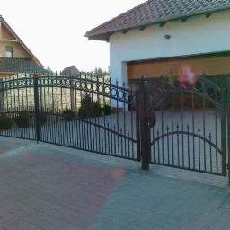 Kuta brama wjazdowa i furtka z ozdobnymi elementami, zamontowana przed domem z kostki brukowej, widok z zewnątrz posesji.