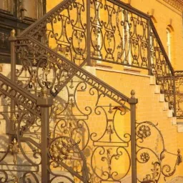 Kute, metalowe balustrady schodowe w stylu retro z ozdobnymi motywami roślinnymi i geometrycznymi, na tle ceglanej elewacji budynku.