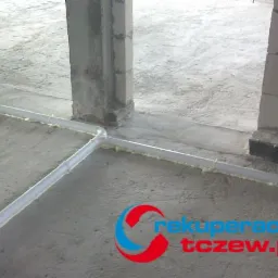 Białe rury instalacji rozprowadzone po surowym betonowym podłożu, zabezpieczone pianką montażową w surowym stanie budynku.