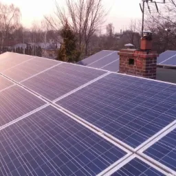 Instalacja fotowoltaiczna na dachu domu z ceglanym kominem w tle, widok z góry. Panele w odcieniach niebieskiego, z delikatną refleksją światła.