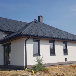 Nowoczesny, parterowy dom szkieletowy z grafitowym dachem i oknami z roletami zewnętrznymi, widok z boku na tle błękitnego nieba i zieleni, teren wokół domu w trakcie zagospodarowywania.