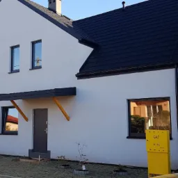 Nowoczesny, biały dom z czarnym dachem i stolarką okienną, z minimalistycznym zadaszeniem nad wejściem wspartym na drewnianych belkach, widok z zewnątrz na świeżo zagospodarowany teren.