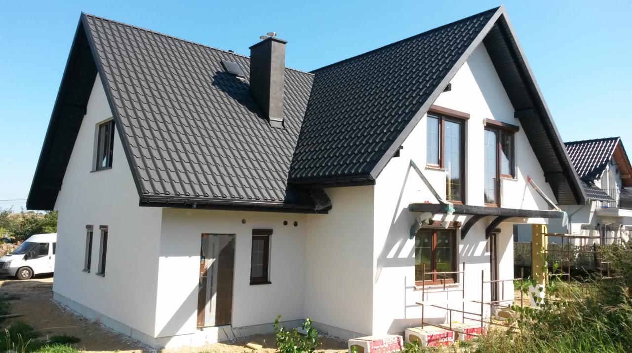 Gotowy budynek z białymi ścianami i ciemnym dachem z dachówki, z widocznym kominem, z oknami z brązowymi ramami, z zewnątrz widać rusztowanie i materiały budowlane.