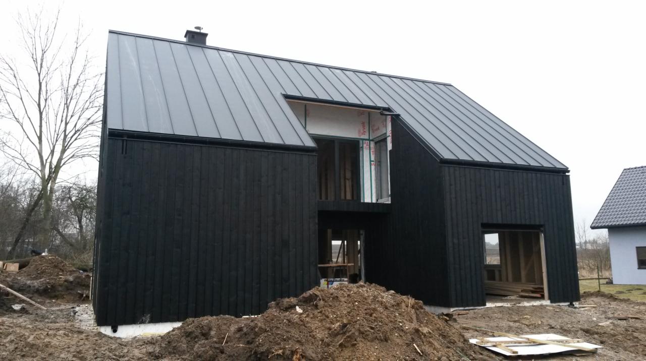 Surowy czarny budynek z drewnianą elewacją i metalowym dachem w trakcie budowy, z widocznymi elementami konstrukcyjnymi i stertą ziemi przed wejściem.