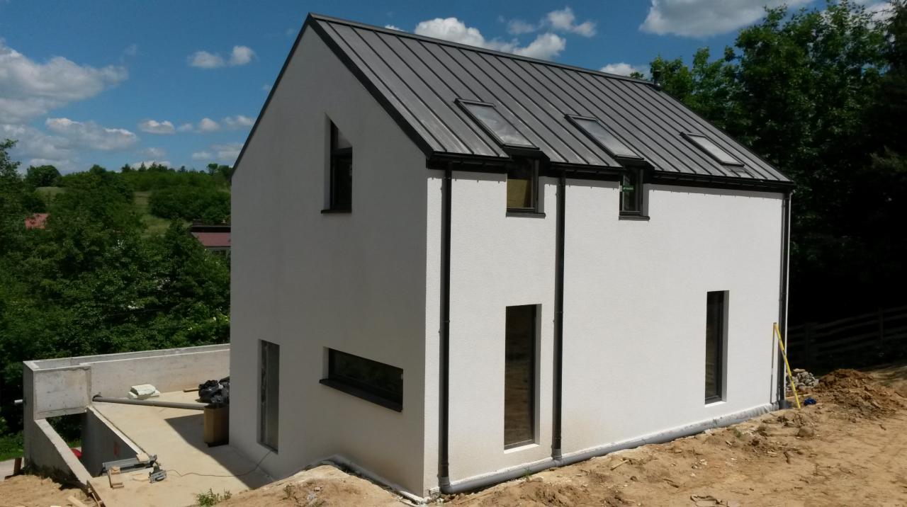 Nowoczesny, biały budynek z szarą blachą na dachu i trzema oknami dachowymi, widoczny w surowym otoczeniu z betonową konstrukcją z boku i stertą ziemi, częściowo ukończony.