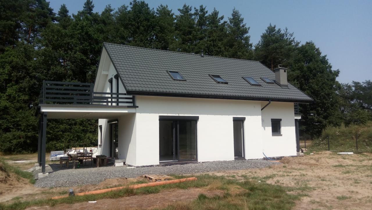 Nowoczesny, biały dom z szarą dachówką i balkonem, otoczony zielenią, widoczne okna dachowe i stolarka okienna w kolorze antracytowym, teren wokół domu w trakcie zagospodarowywania.
