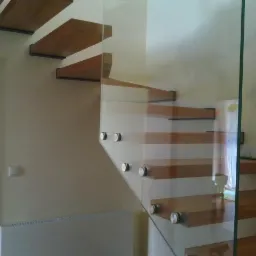 Nowoczesne schody z drewnianymi stopniami, mocowane do ściany z balustradą ze szkła, widoczne metalowe okucia i grzejnik na ścianie.