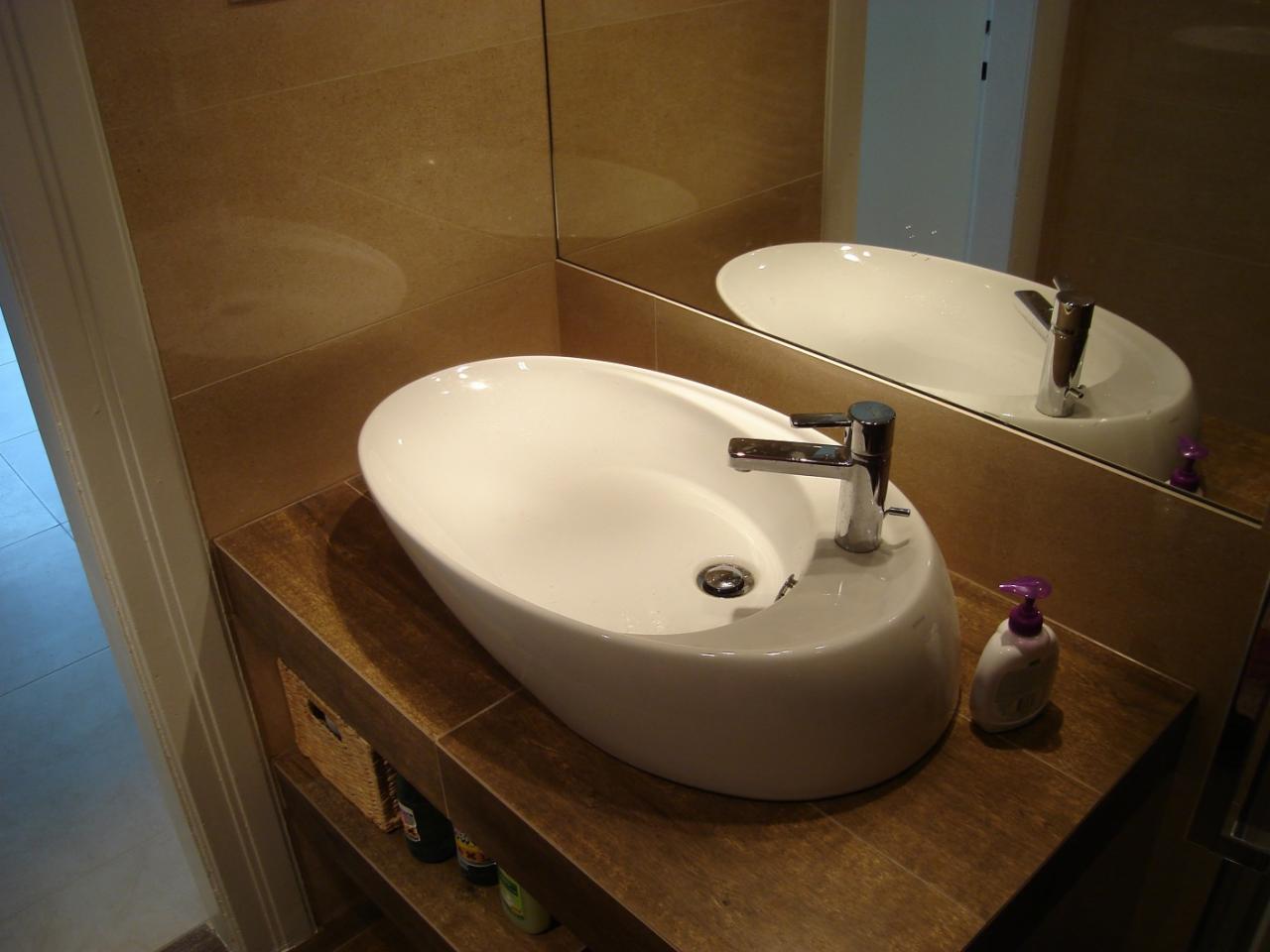 Nowoczesna, owalna umywalka nablatowa z chromowaną baterią, stojąca na drewnianym blacie w łazience wykończonej w odcieniach beżu i brązu, z widoczną butelką mydła w płynie.