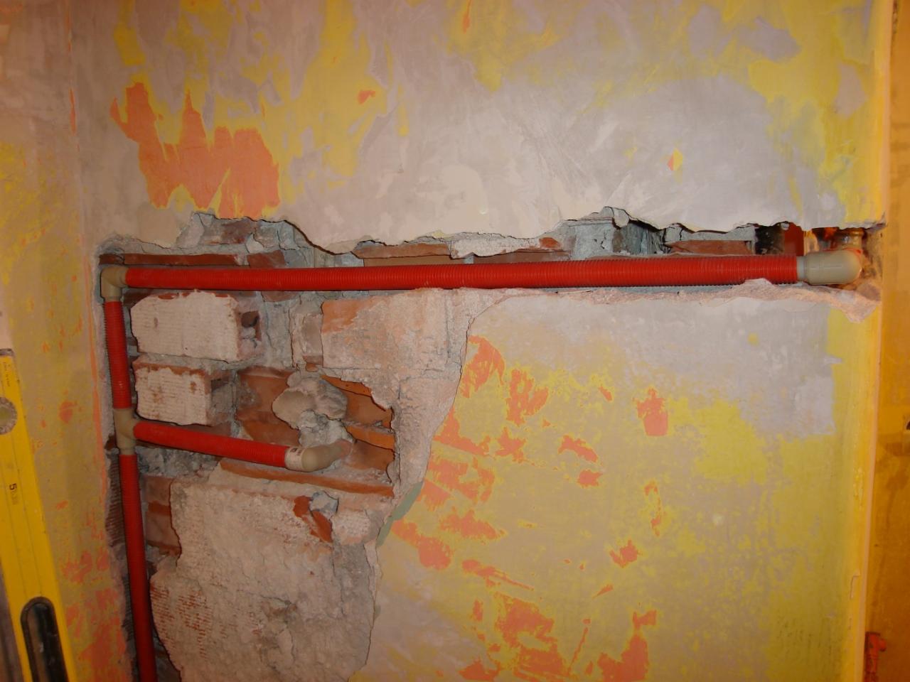 Czerwone rury instalacji wodnej, częściowo schowane w bruździe wyciętej w starej, częściowo otynkowanej ścianie z cegły. Widoczne ślady po wcześniejszych warstwach farby na ścianie i poziomica przy...