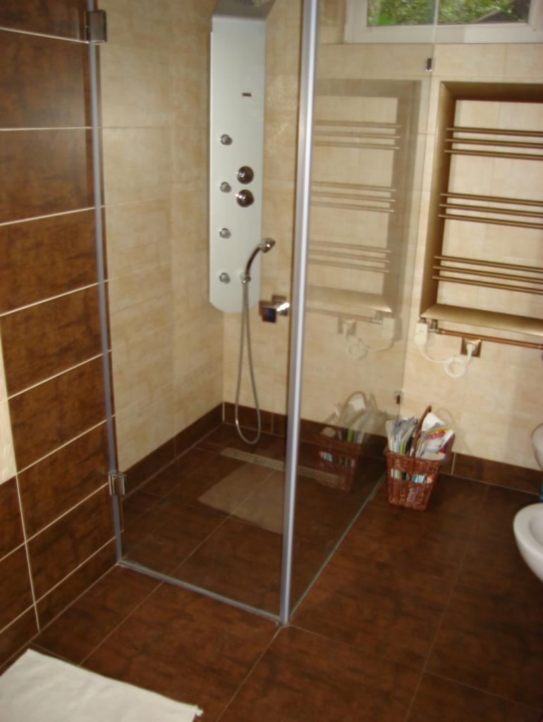 Wykończona łazienka z kabiną prysznicową typu walk-in, panelem natryskowym z hydromasażem i grzejnikiem drabinkowym, wykończona brązowymi i beżowymi płytkami, widoczny kosz z gazetami.