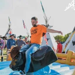 Uśmiechnięty mężczyzna w pomarańczowej koszulce i dżinsach, w kapeluszu kowbojskim, próbuje utrzymać się na mechanicznym byku podczas pikniku w stylu western, w tle tipi i osoby w kowbojskich...