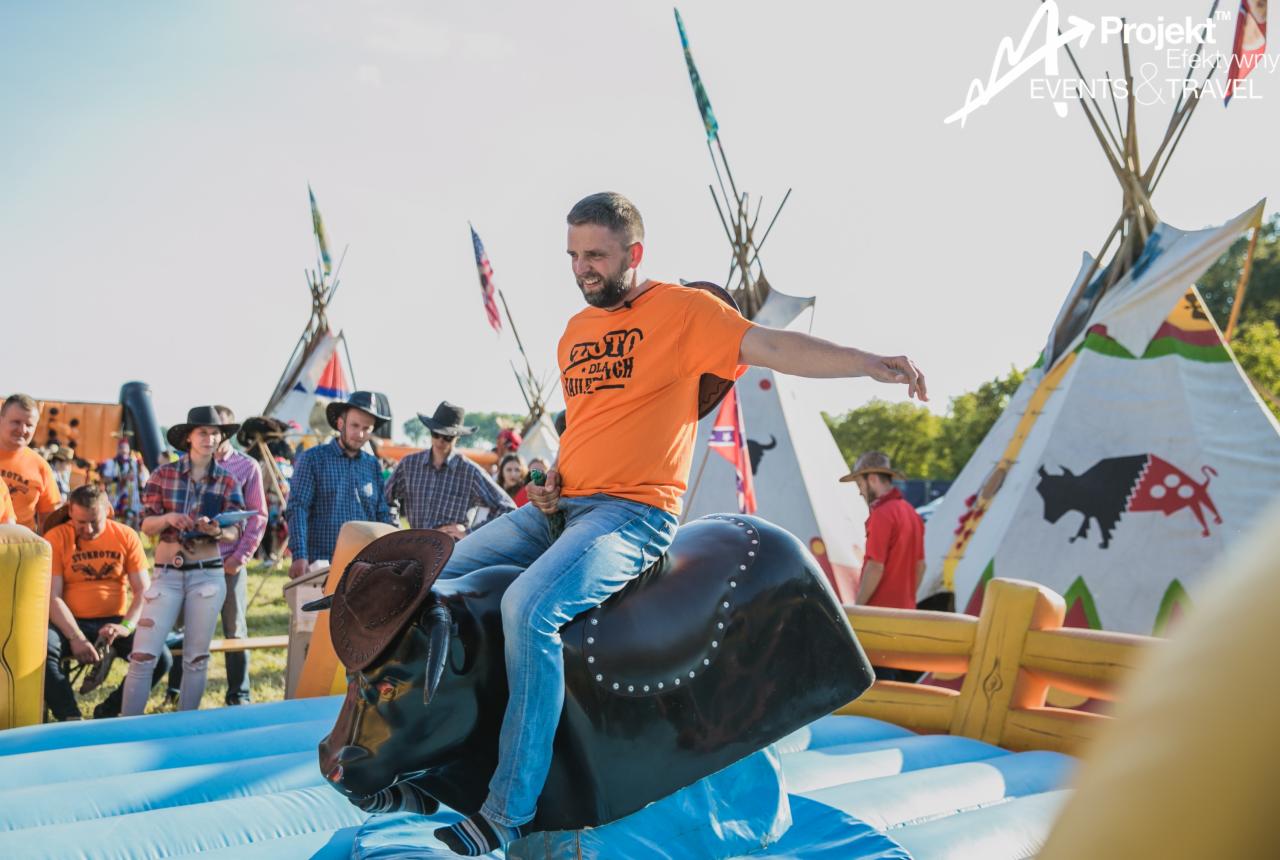 Uśmiechnięty mężczyzna w pomarańczowej koszulce i dżinsach, w kapeluszu kowbojskim, próbuje utrzymać się na mechanicznym byku podczas pikniku w stylu western, w tle tipi i osoby w kowbojskich...