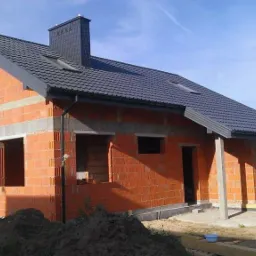 Budowa domu jednorodzinnego w stanie surowym zamkniętym z dachem pokrytym blachodachówką, widoczne pustaki Porotherm oraz komin z czarnej dachówki.