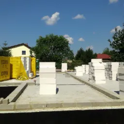 Przygotowany fundament z wyeksponowanymi paletami bloczków Ytong w żółtej folii ochronnej, czekających na dalsze prace murarskie, w tle domy jednorodzinne pod błękitnym niebem.