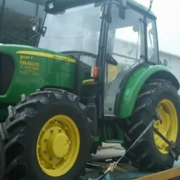 Zielony ciągnik rolniczy John Deere 5065E zabezpieczony pasami transportowymi na żółtej platformie lawety, na tle budynku z bramami garażowymi i zaparkowanym samochodem.