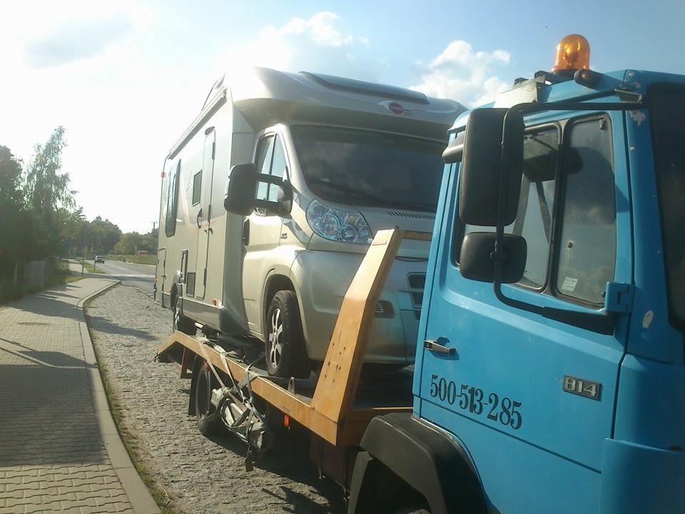 Błękitna ciężarówka z żółtym wysięgnikiem przewozi kampera na platformie, widoczny numer telefonu na drzwiach kabiny.