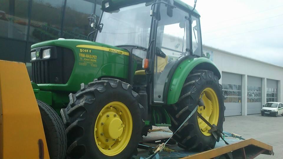 Zielony ciągnik rolniczy John Deere 5065E zabezpieczony pasami transportowymi na żółtej platformie lawety, na tle budynku z bramami garażowymi i zaparkowanym samochodem.