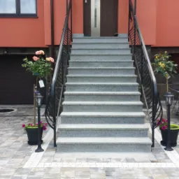 Schody wejściowe z szarej kostki brukowej prowadzące do domu o pomarańczowej elewacji, z dekoracyjnymi roślinami w donicach i oświetleniem, oraz fragmentem tarasu z meblami ogrodowymi.