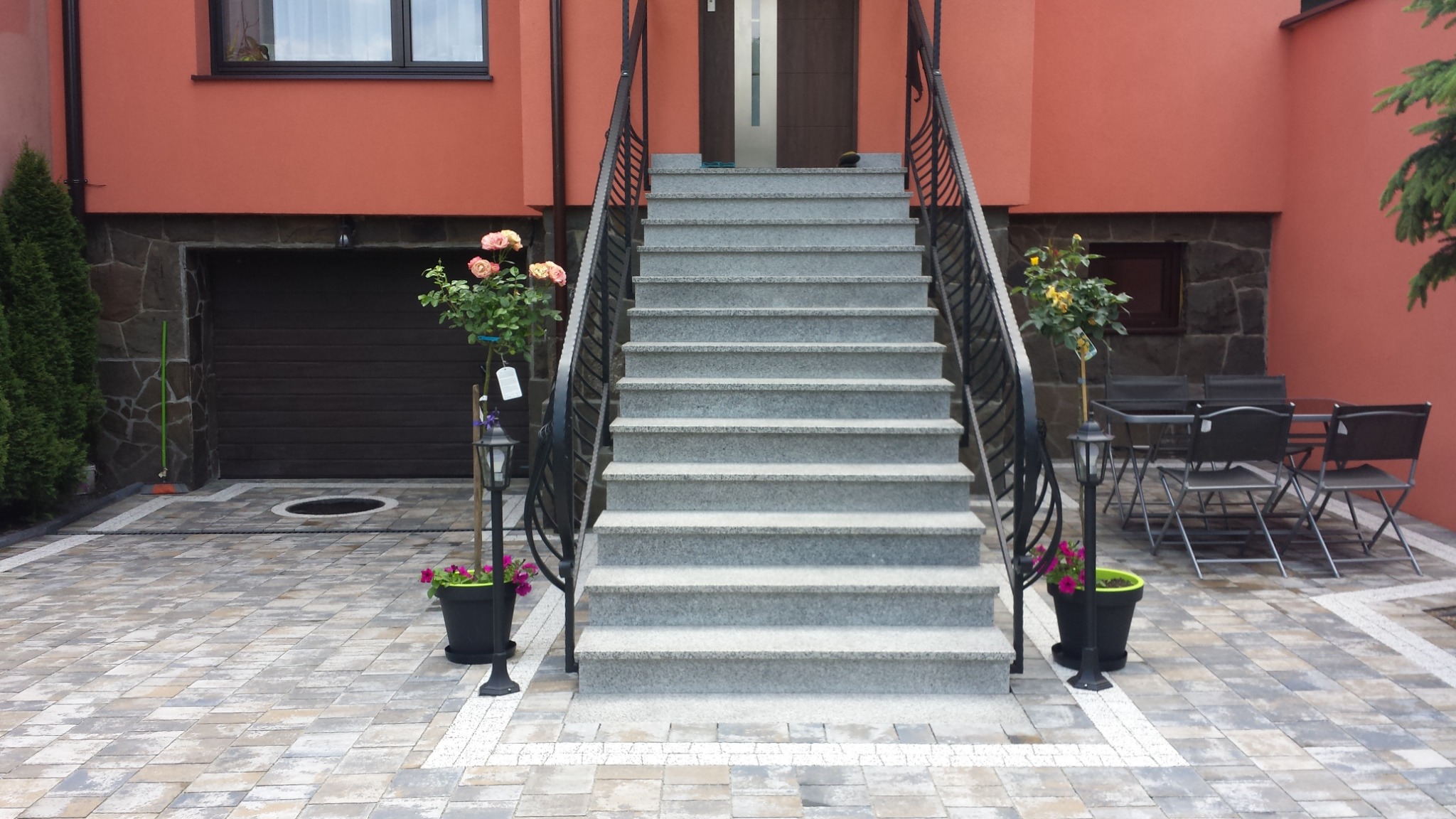 Schody wejściowe z szarej kostki brukowej prowadzące do domu o pomarańczowej elewacji, z dekoracyjnymi roślinami w donicach i oświetleniem, oraz fragmentem tarasu z meblami ogrodowymi.
