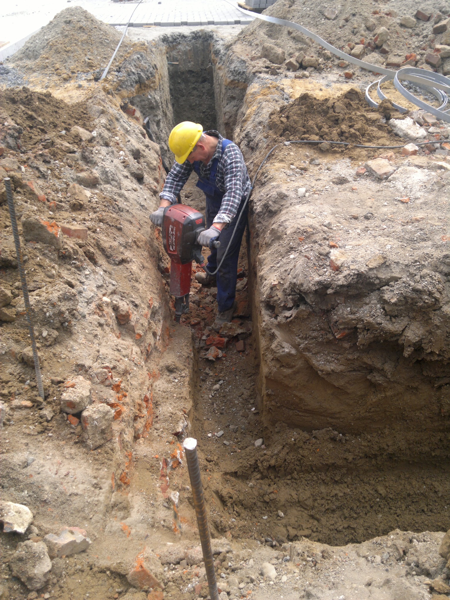 Pracownik w kasku i rękawicach używa młota pneumatycznego w wąskim wykopie, widoczne ściany wykopu z fragmentami cegieł i ziemi.