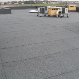 Płaski dach pokryty papą termozgrzewalną z widocznymi klimatyzatorami i kominami wentylacyjnymi na tle zabudowań.