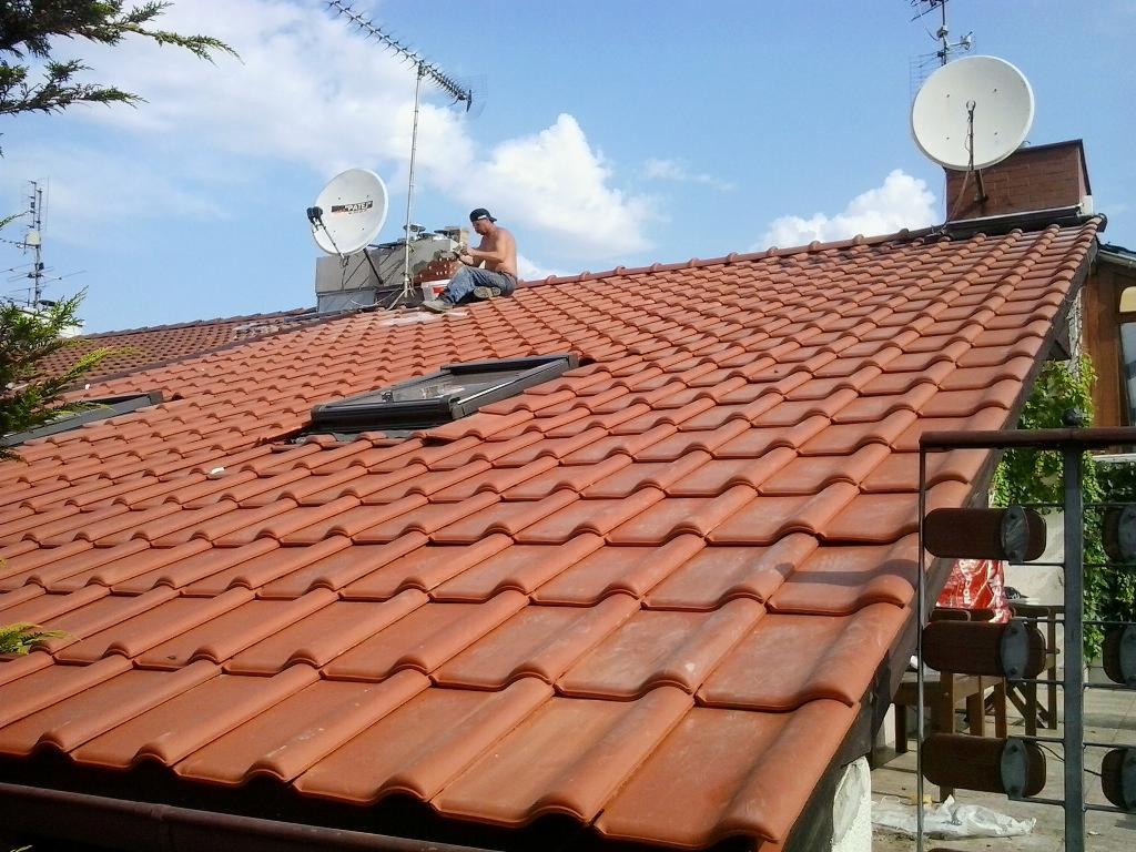 Czerwona dachówka ceramiczna z oknami dachowymi, na dachu siedzi dekarz w czapce, obok anteny satelitarne i telewizyjne, błękitne niebo z chmurami w tle.
