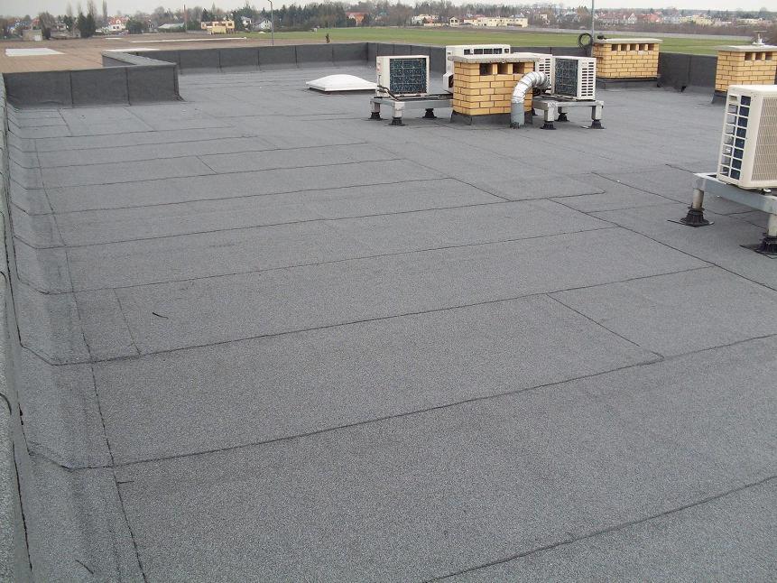 Płaski dach pokryty papą termozgrzewalną z widocznymi klimatyzatorami i kominami wentylacyjnymi na tle zabudowań.