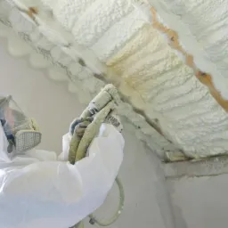 Pracownik w kombinezonie natryskuje pianę PUR na poddasze, tworząc izolację. Widoczny pistolet natryskowy i drewniane elementy konstrukcyjne pokryte pianą. Jasne oświetlenie.