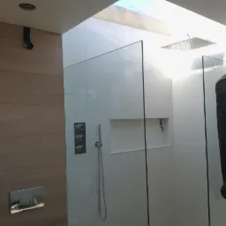 Nowoczesna kabina prysznicowa z białymi płytkami, szklanymi ściankami i deszczownicą. Widoczne elementy armatury i wnęka w ścianie. Na szybie wisi pokrowiec na ubranie.